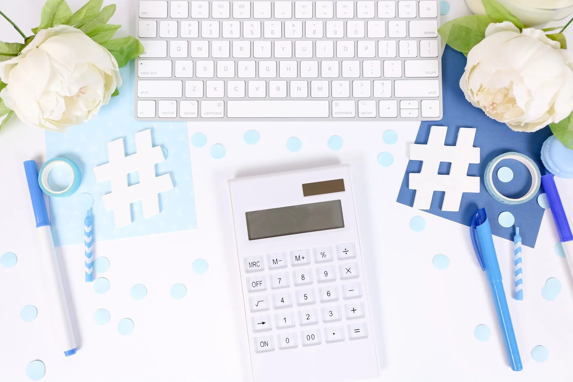 A white calculator and hashtag cutouts on a desk beside a keyboard and white flowers
