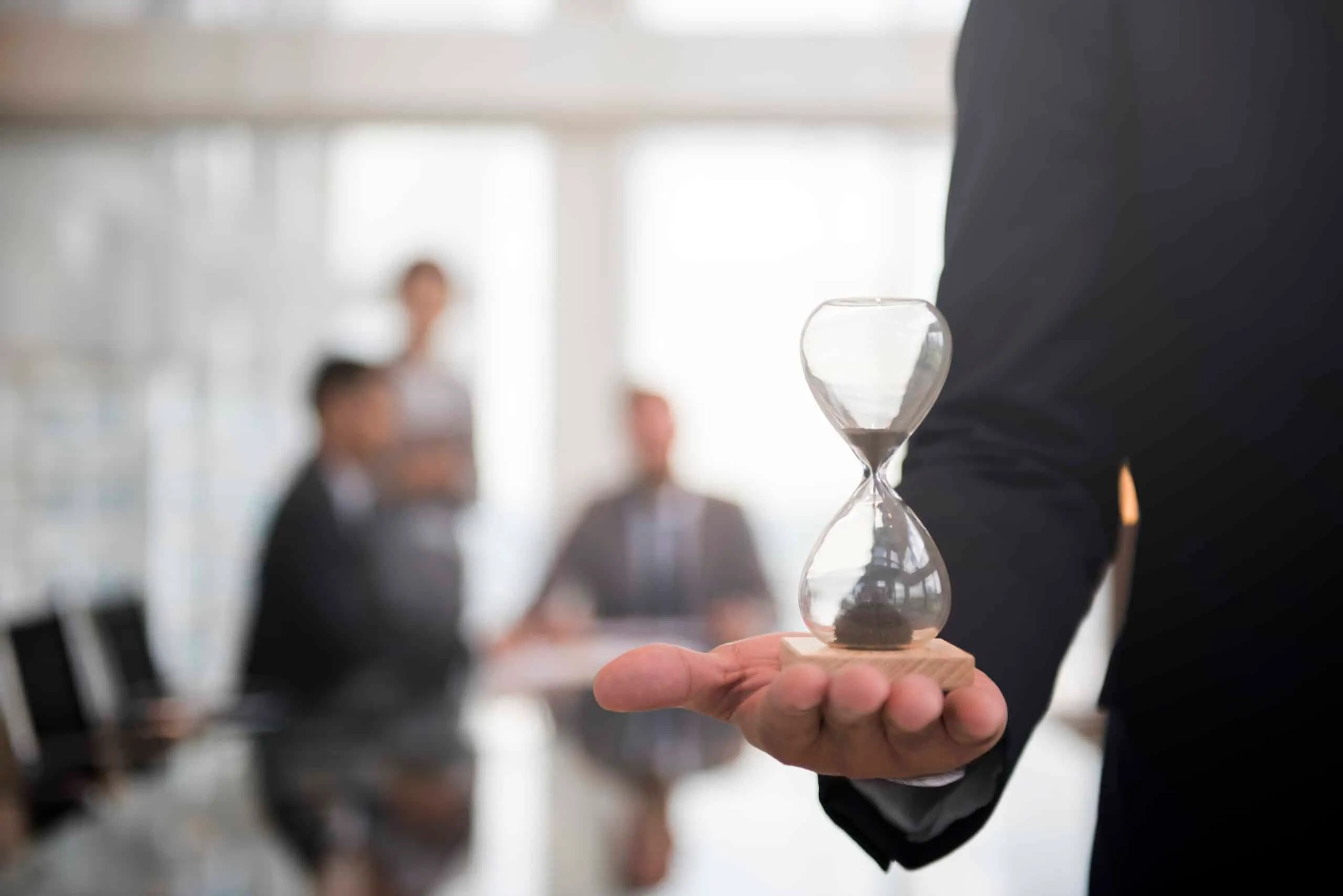 A hand holding a small hourglass, with blurred office colleagues in the background.