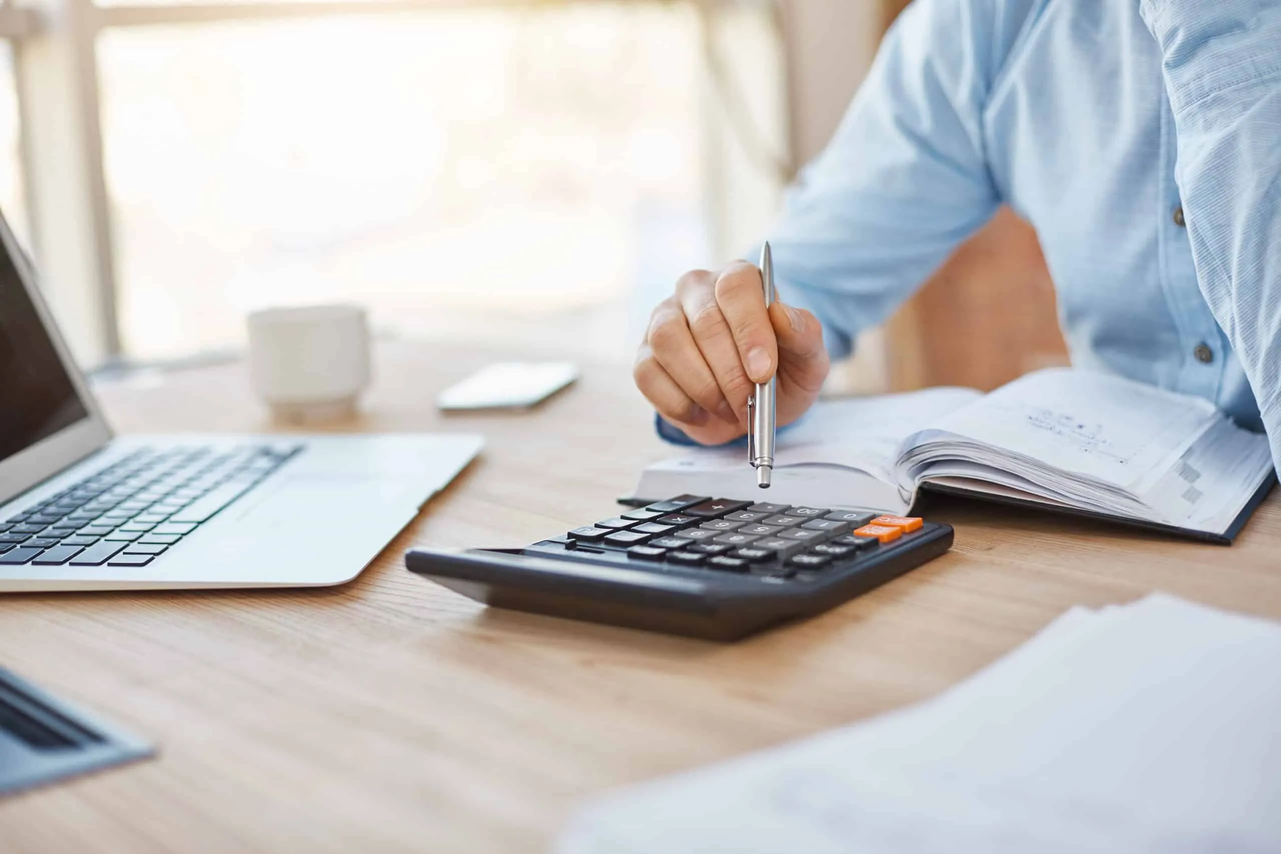 A hand holding a pen over an open notebook next to a calculator and laptop.