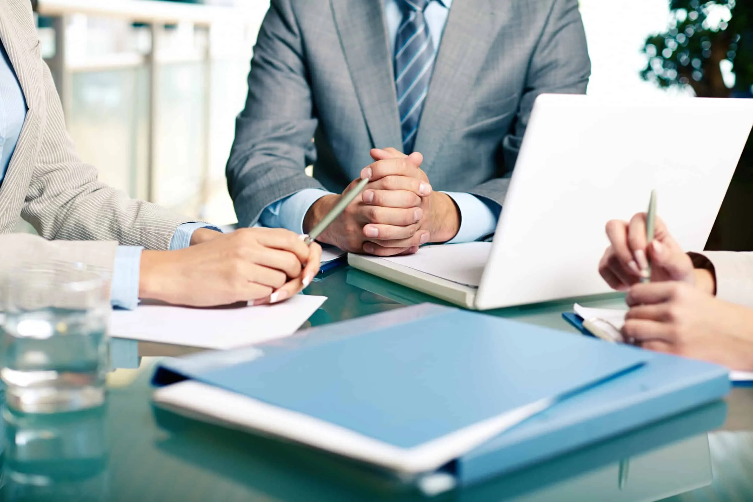 Close-up of hands and pens around a glass meeting table with a laptop and folders.