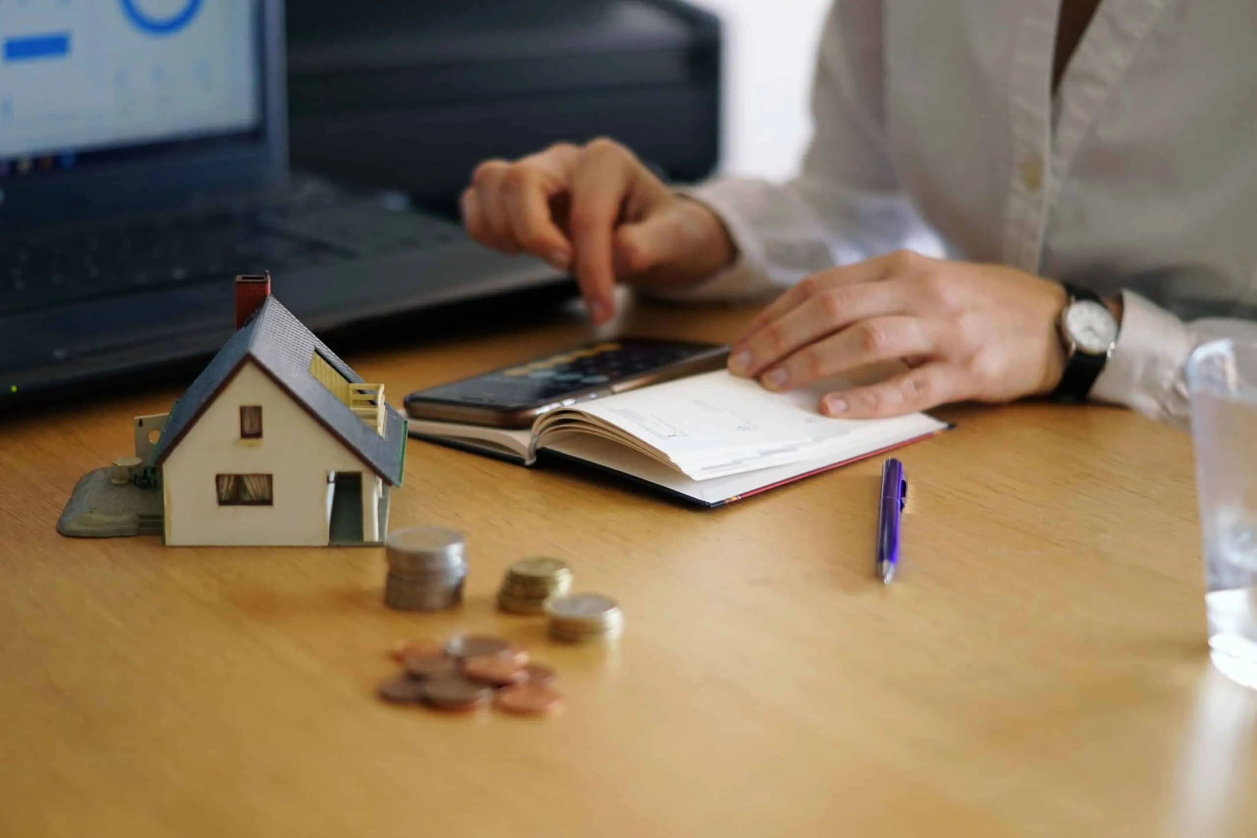 A hand beside a model house and stacked coins, with a notebook and laptop nearby.