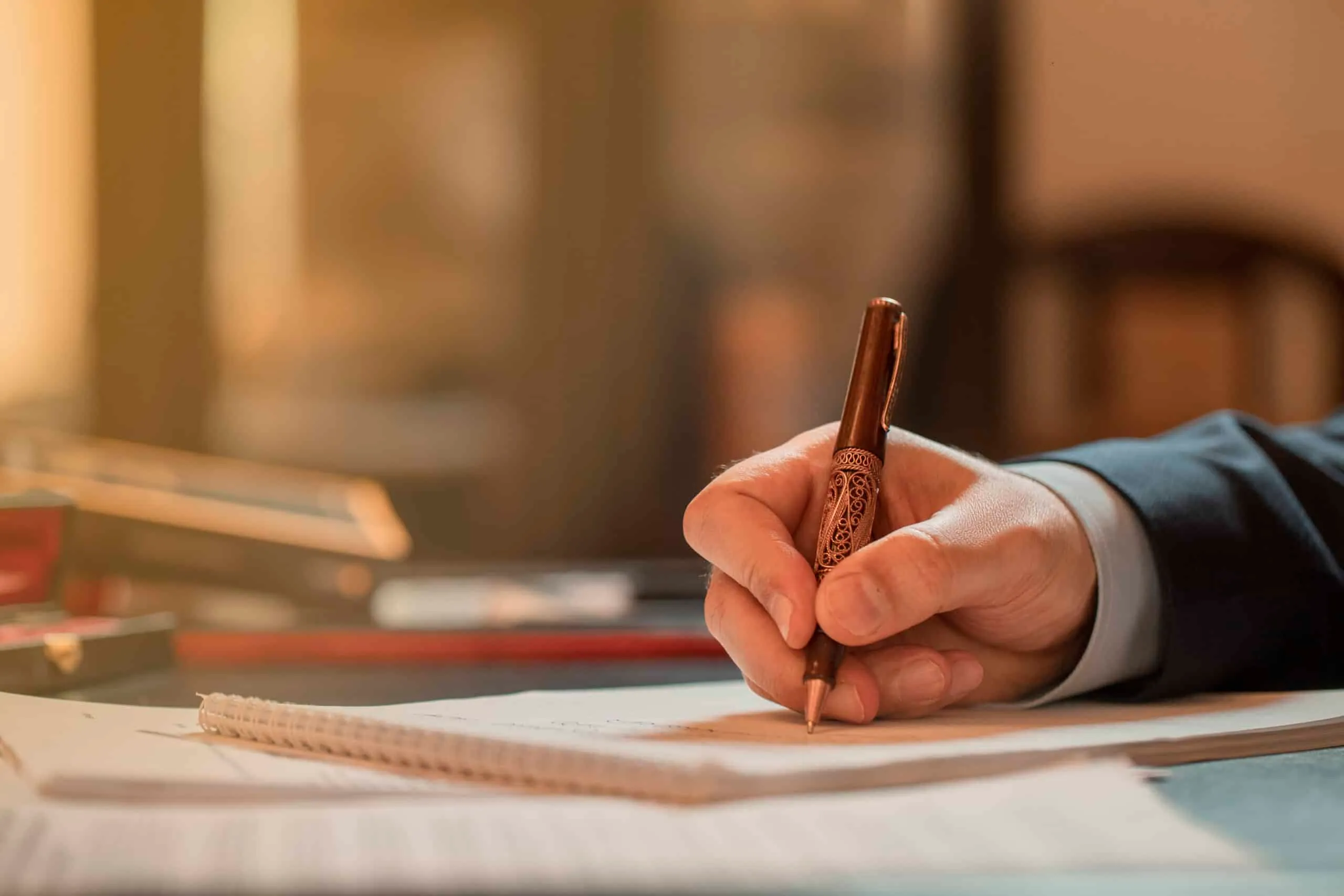 Close-up of a hand in a suit sleeve signing a document with an ornate pen.