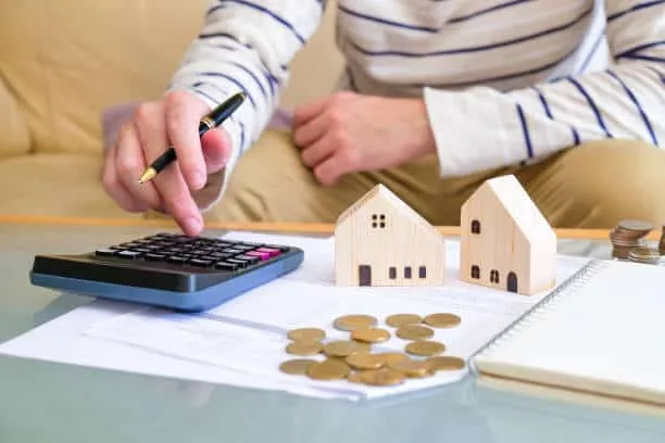Hand pressing calculator buttons beside a pen, coins and two small wooden house models