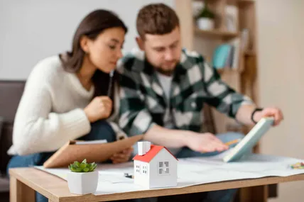 Blurred couple on a sofa reviewing documents behind a small toy house and plant