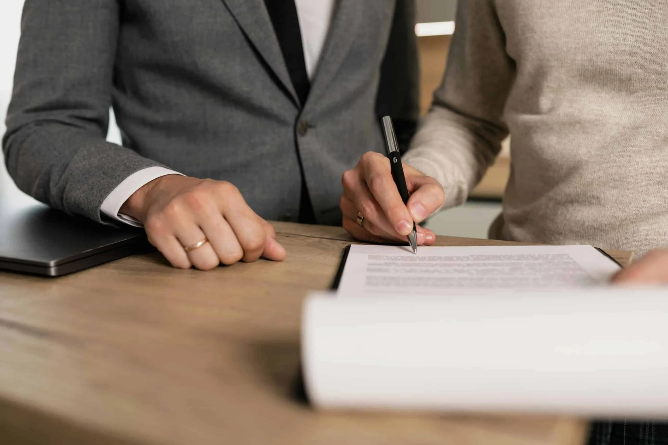 Hand signing a document with a pen beside a laptop on a wooden desk