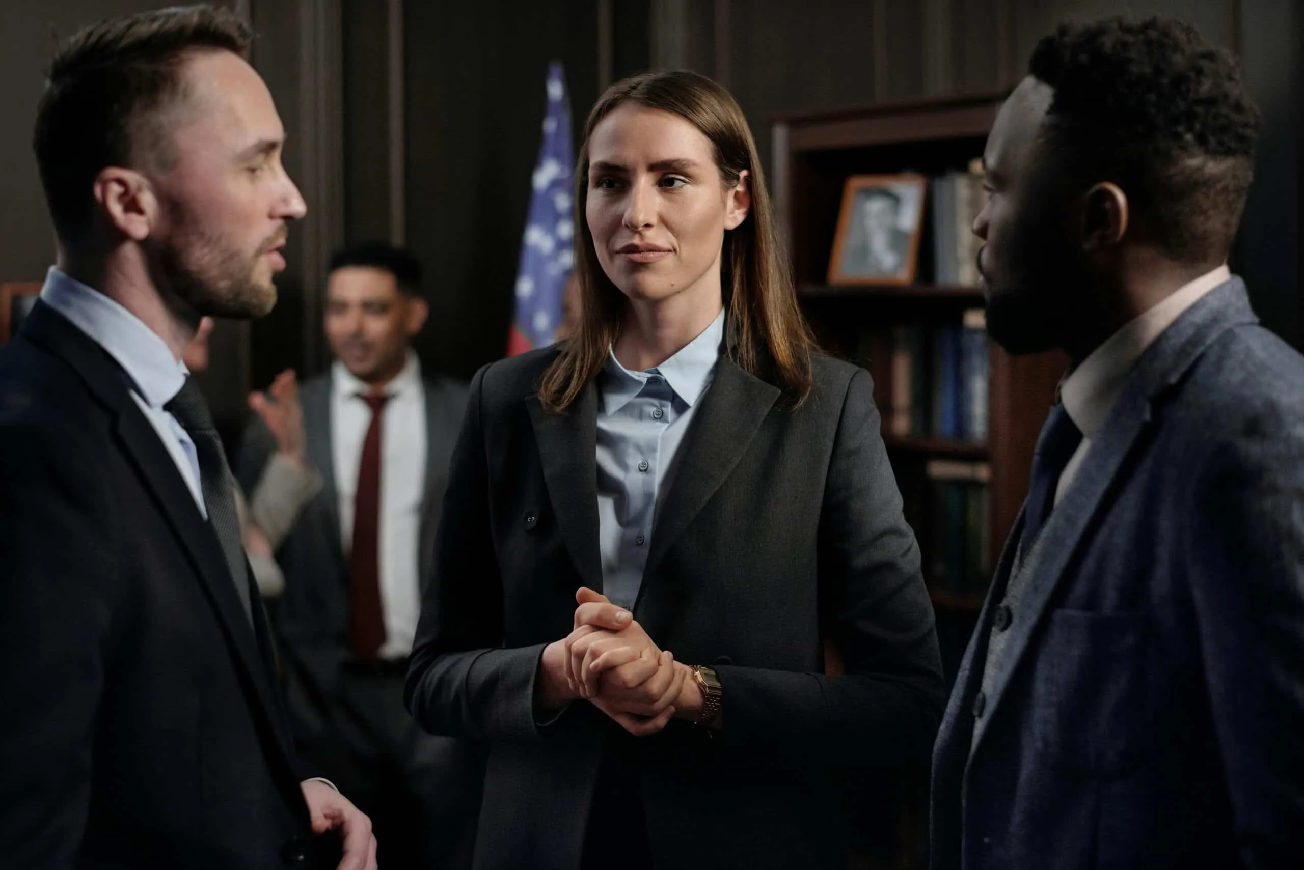 Group of people in business suits talking in an office with a flag and bookshelf