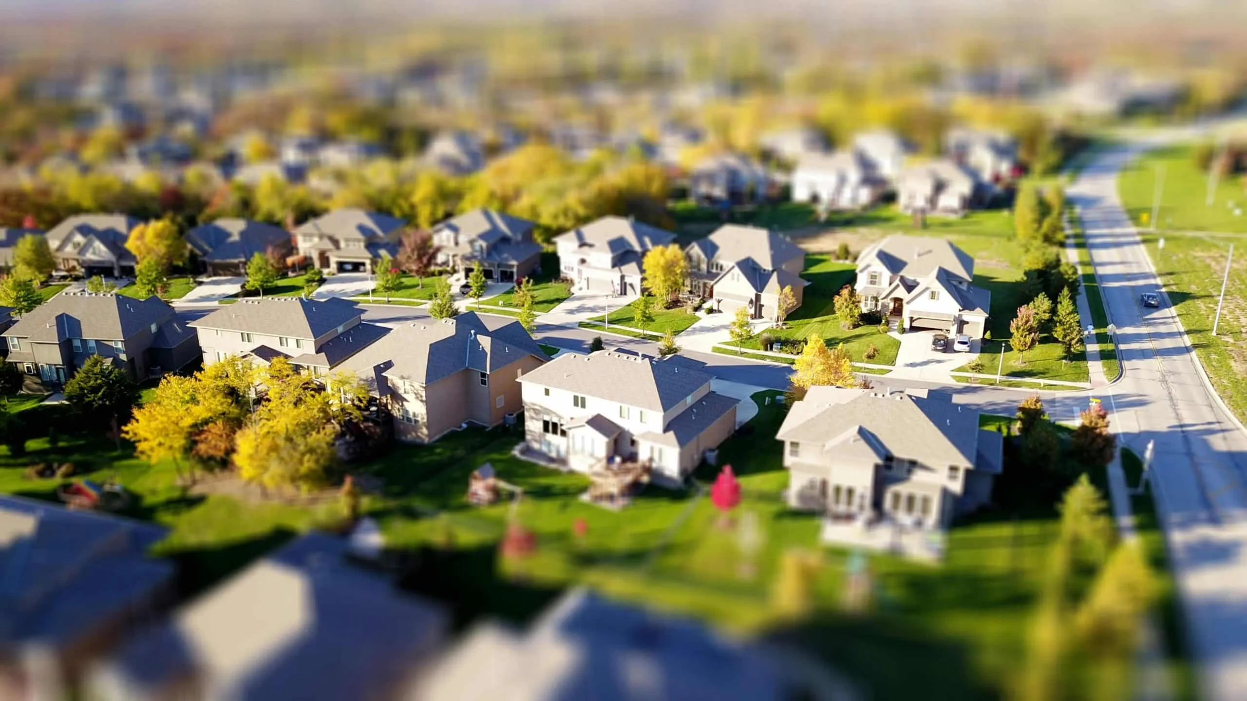 Aerial tilt-shift view of a suburban street lined with houses, lawns and trees