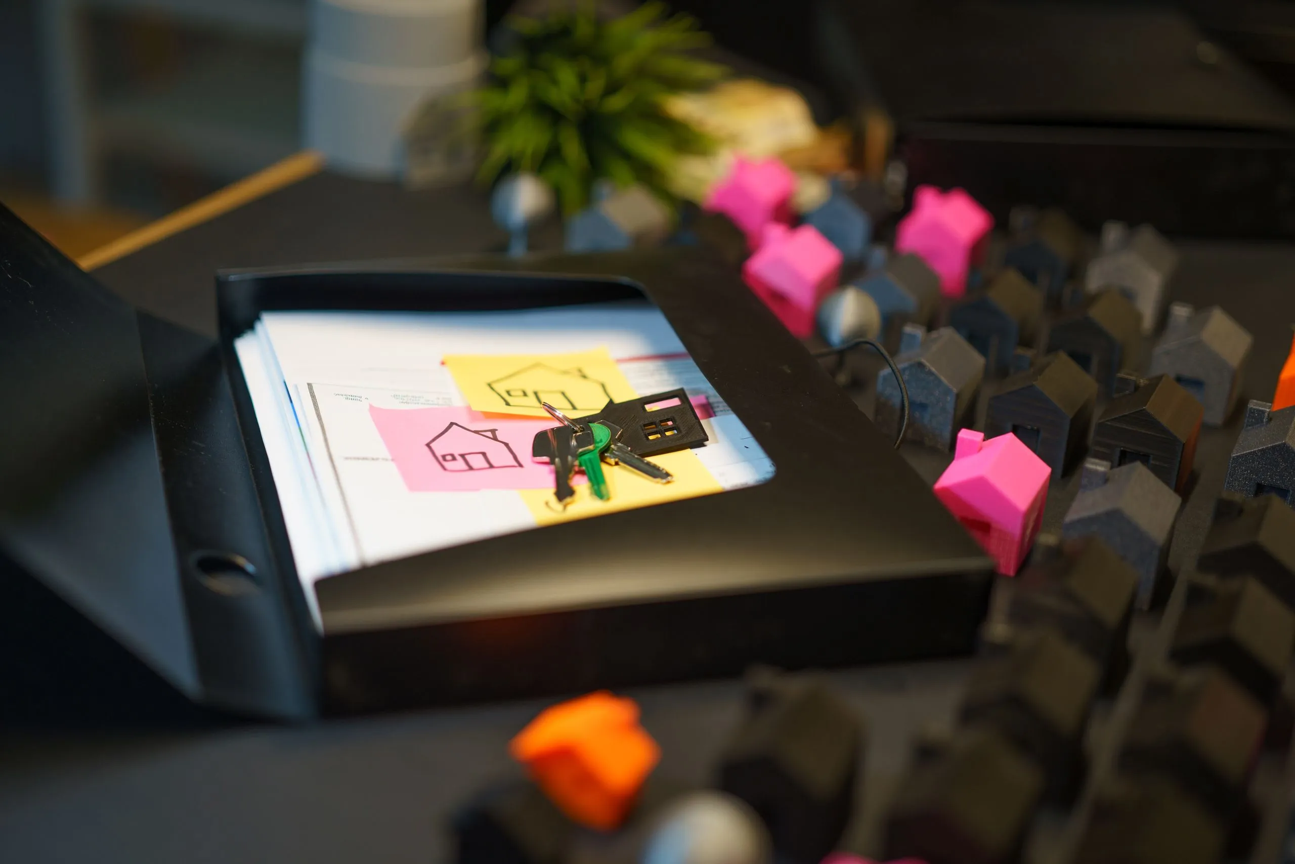 Keys and hand-drawn house sketches on a stack of paperwork, with miniature house models arranged on a desk in the background.