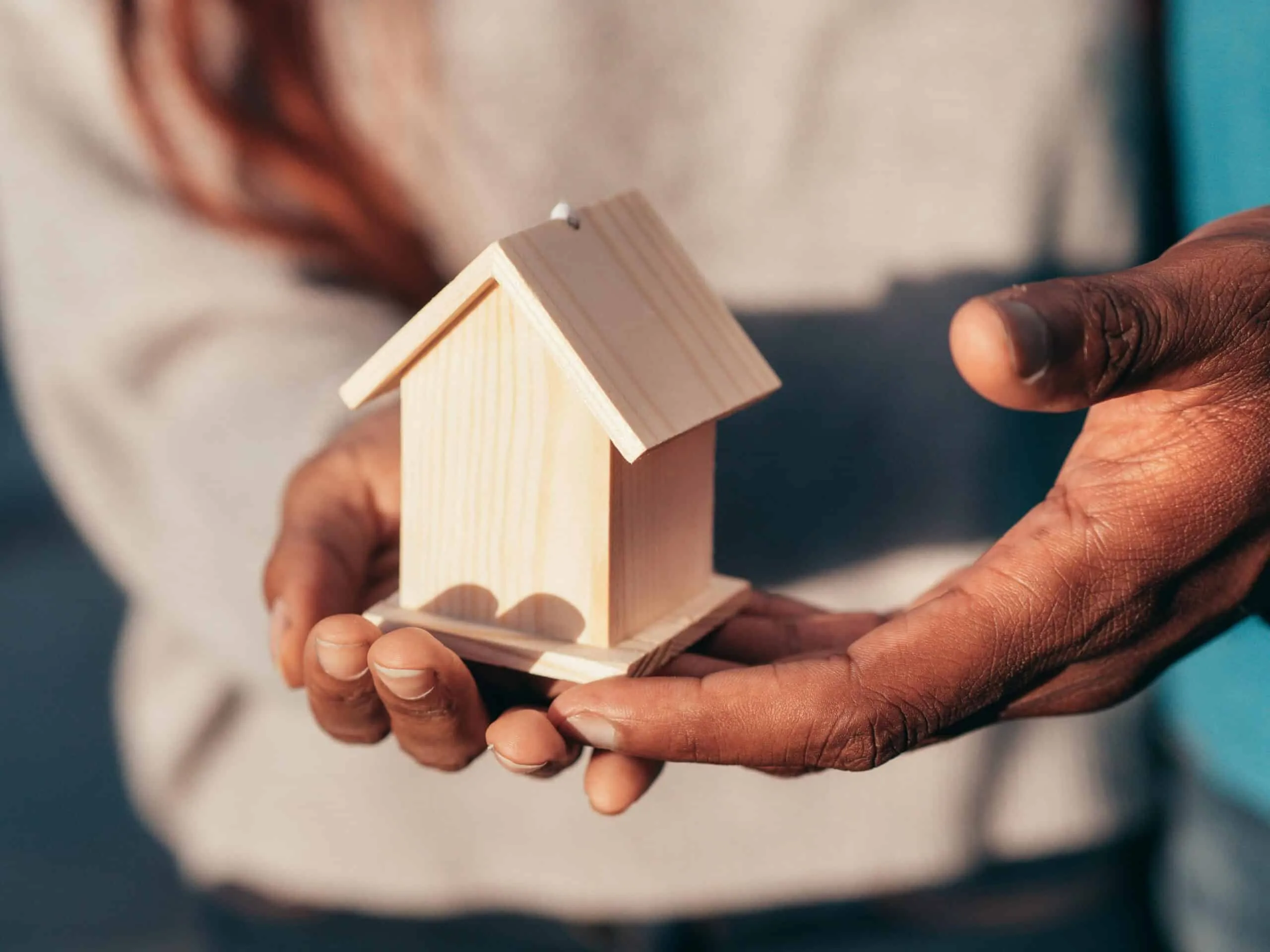Two hands cradle a small wooden model house in sunlight