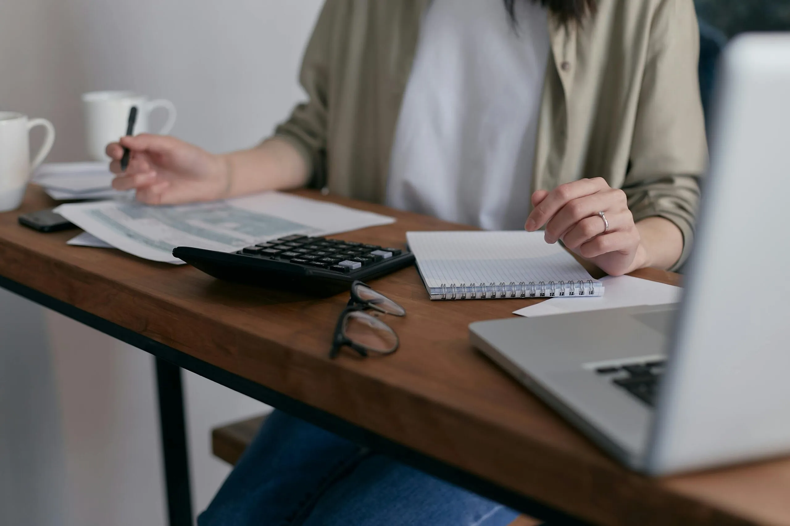 A person at a desk with a calculator, notebook, glasses and an open laptop