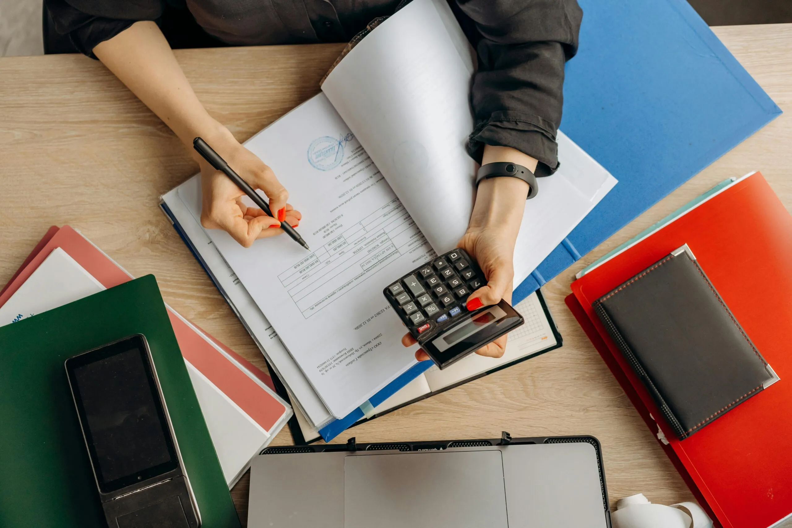 Overhead view of hands holding a pen and calculator beside folders and a signed document