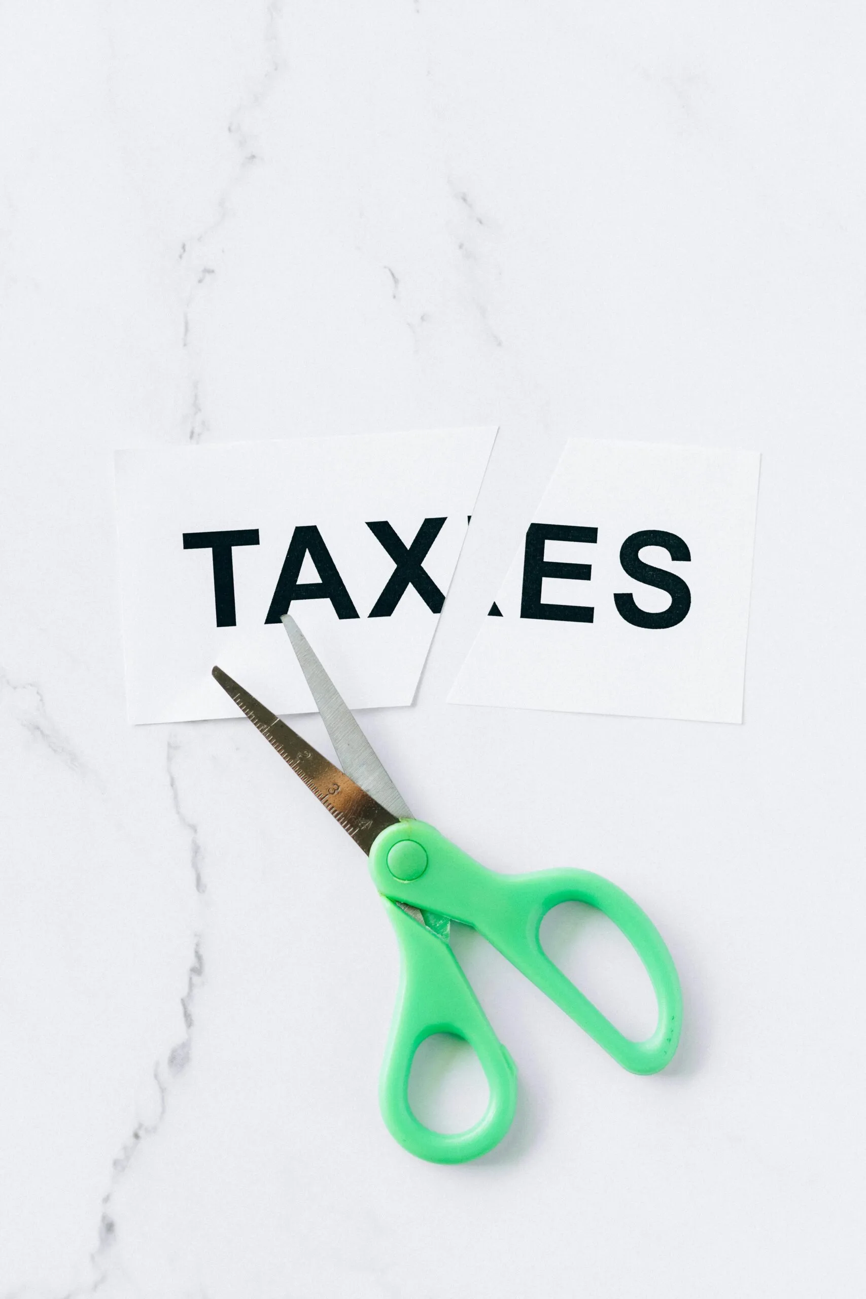 Green-handled scissors cutting a paper sign reading TAXES on a white marble surface