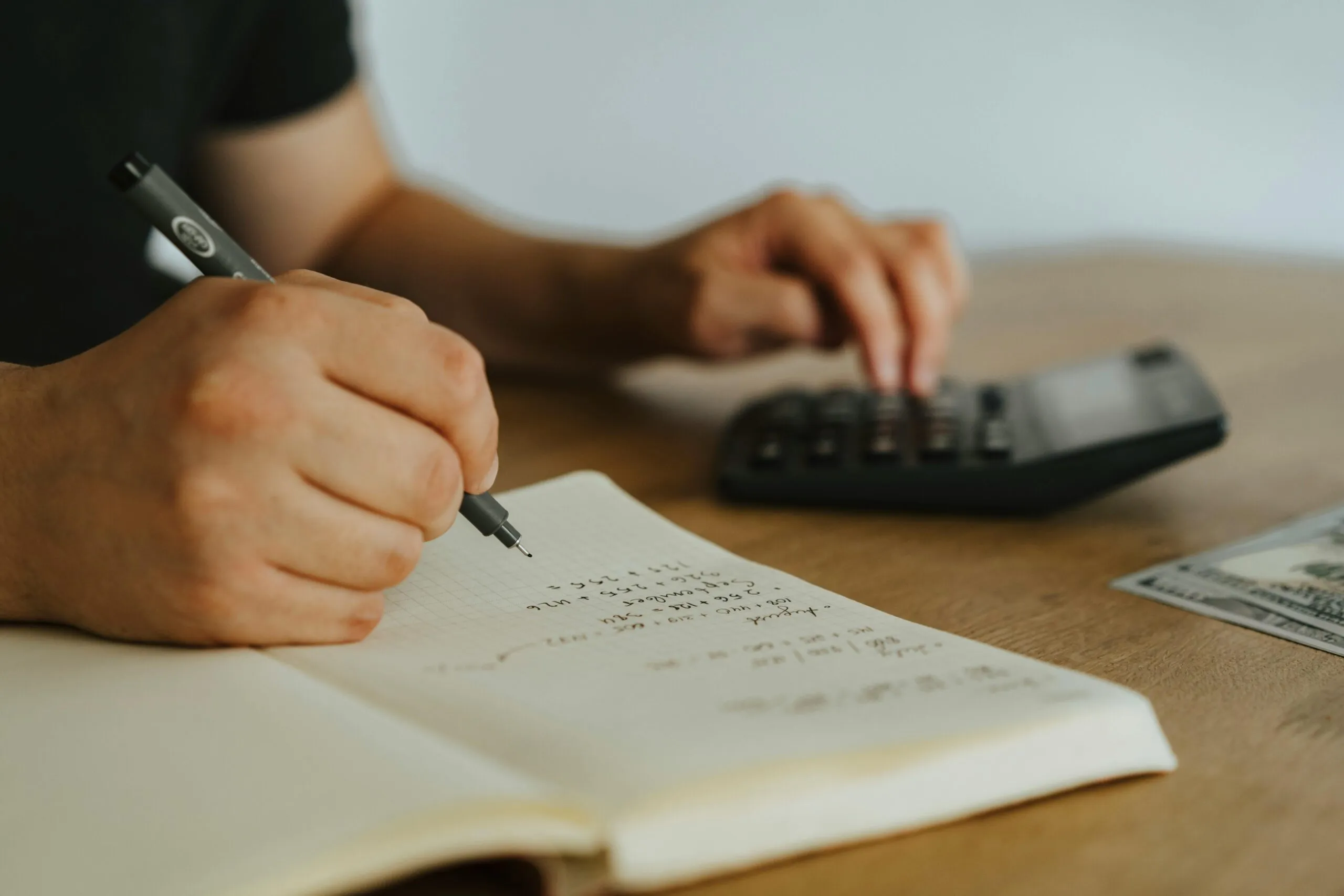 Close-up of a hand writing calculations in a notebook beside a calculator and cash