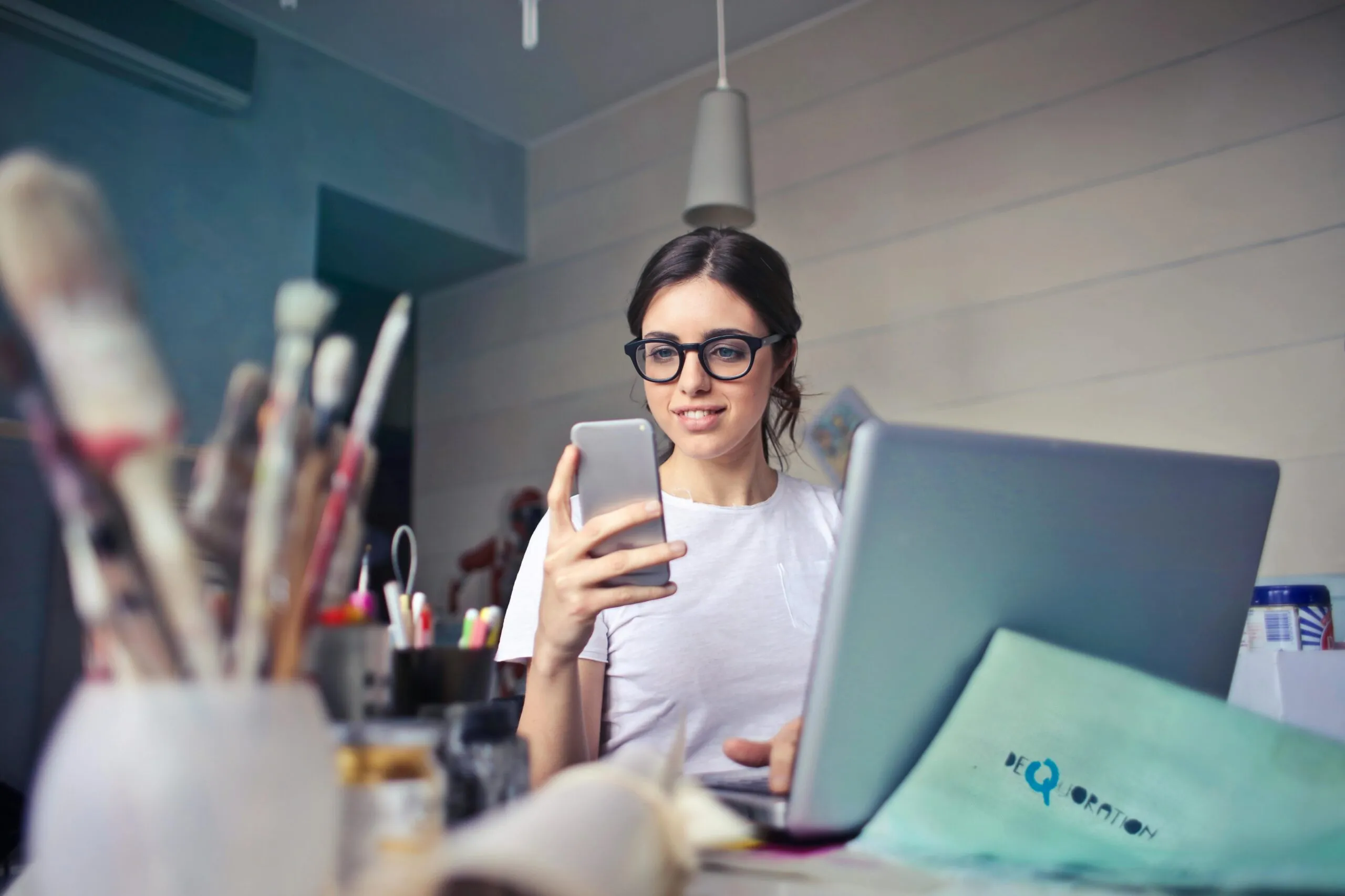 Woman wearing glasses smiles while holding a smartphone at a desk with a laptop