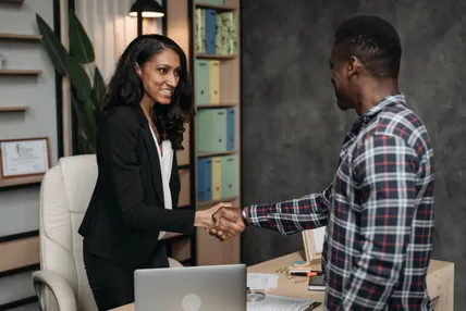 A businesswoman and a client shaking hands in an office, with folders and a laptop on the desk beside them.