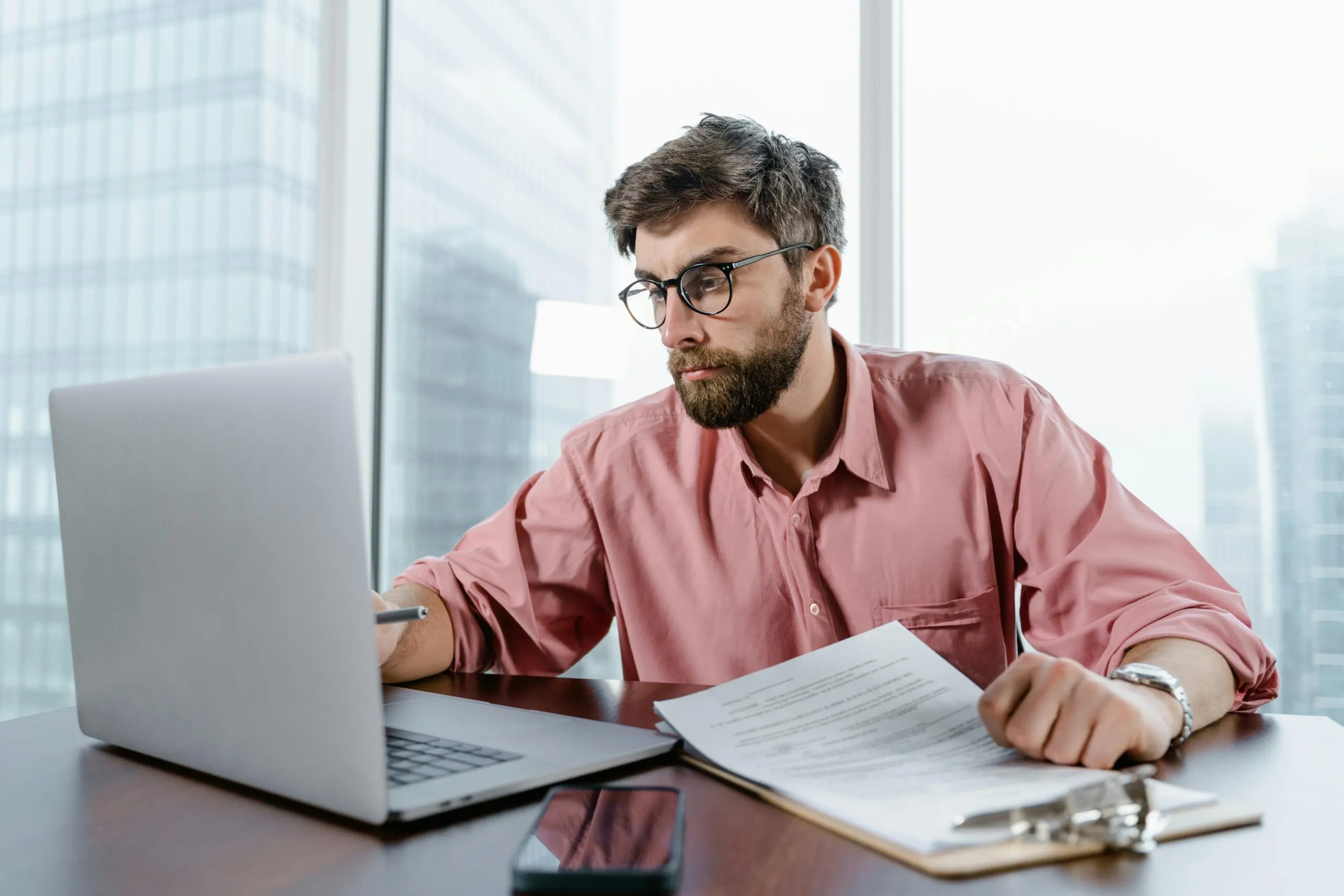 Bearded man in glasses reviewing a document beside an open laptop in an office