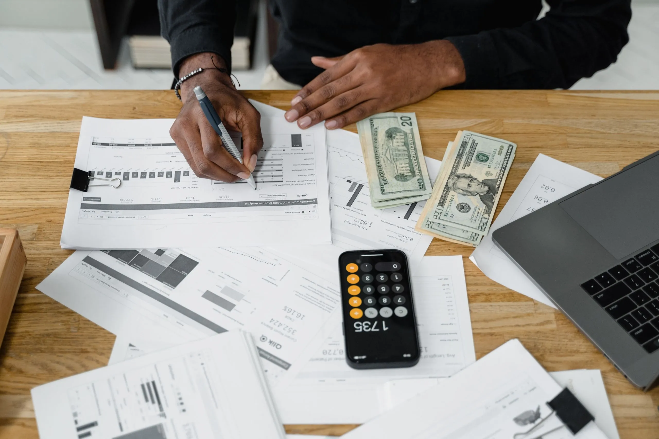 A hand writing on printed financial reports next to a calculator, banknotes and an open laptop.