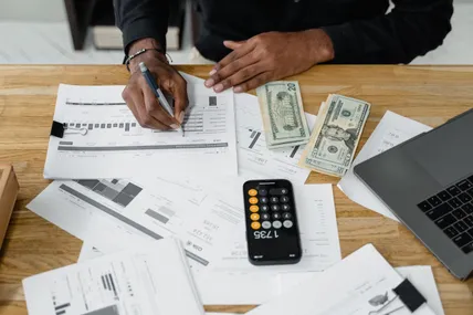 A hand writing on printed financial reports next to a calculator, banknotes and an open laptop.