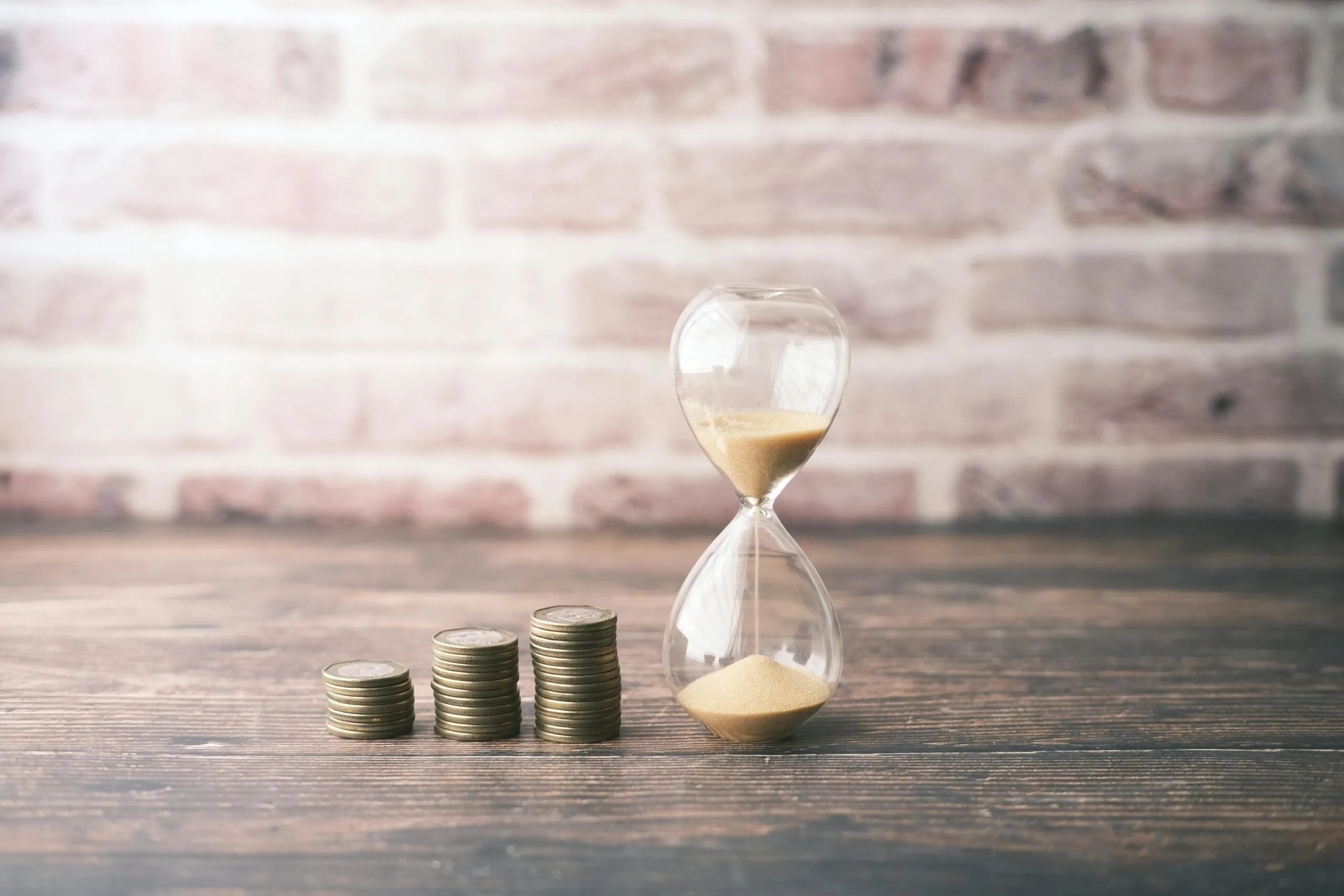 An hourglass beside three ascending stacks of coins on a wooden table
