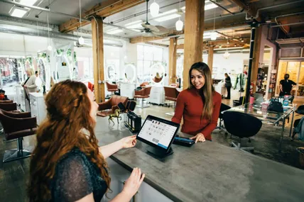 Two women chatting at a salon checkout counter with a point of sale tablet
