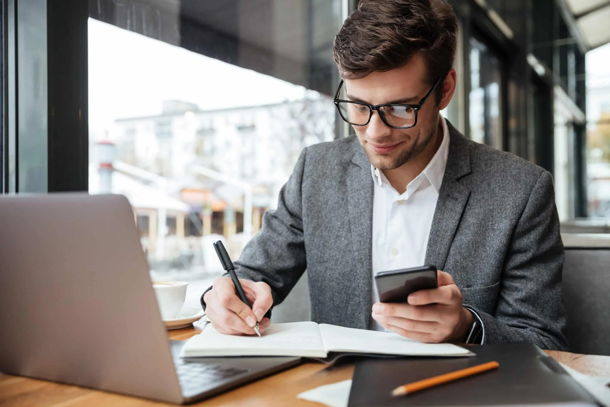 Man in glasses writing in a notebook while holding a phone at a cafe table