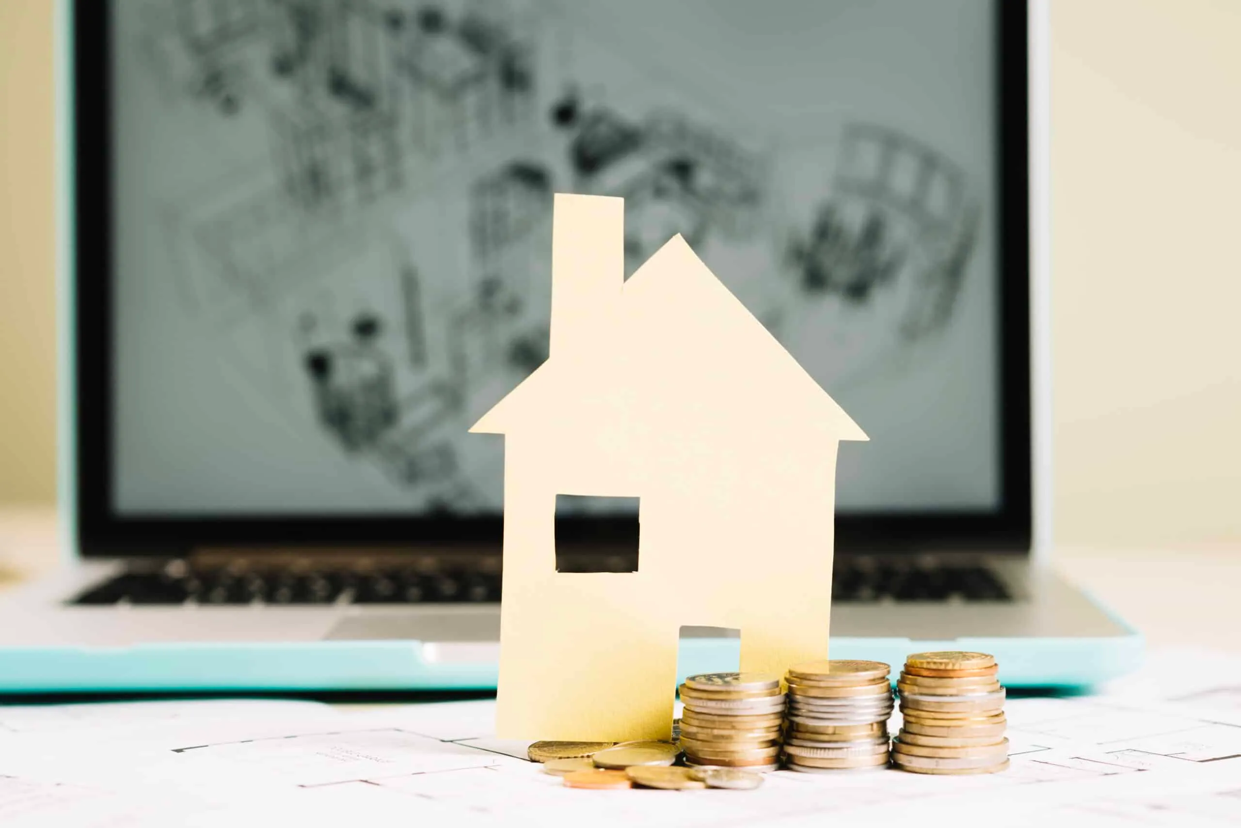 Paper cut-out house standing beside stacked coins in front of a laptop showing house blueprints
